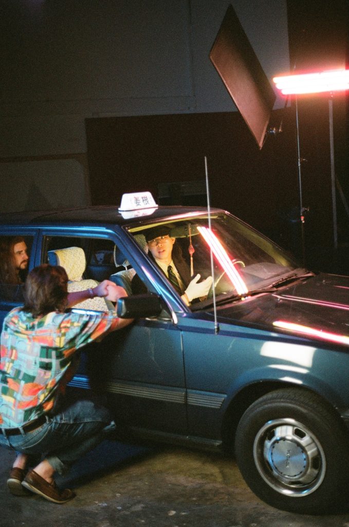 Ginger Root sits inside of a black taxi in the passenger side, while wearing a black suit. Someone crouches to the side hanging on the passenger window. Another person sits in the back seat