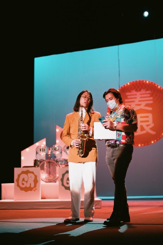 Ginger Root Stands in a white suite with a orange blazer on top. Ginger stands with a Saxophone in hand. Next to him stands a man in a brightly colored outfit, giving an award to Ginger. 