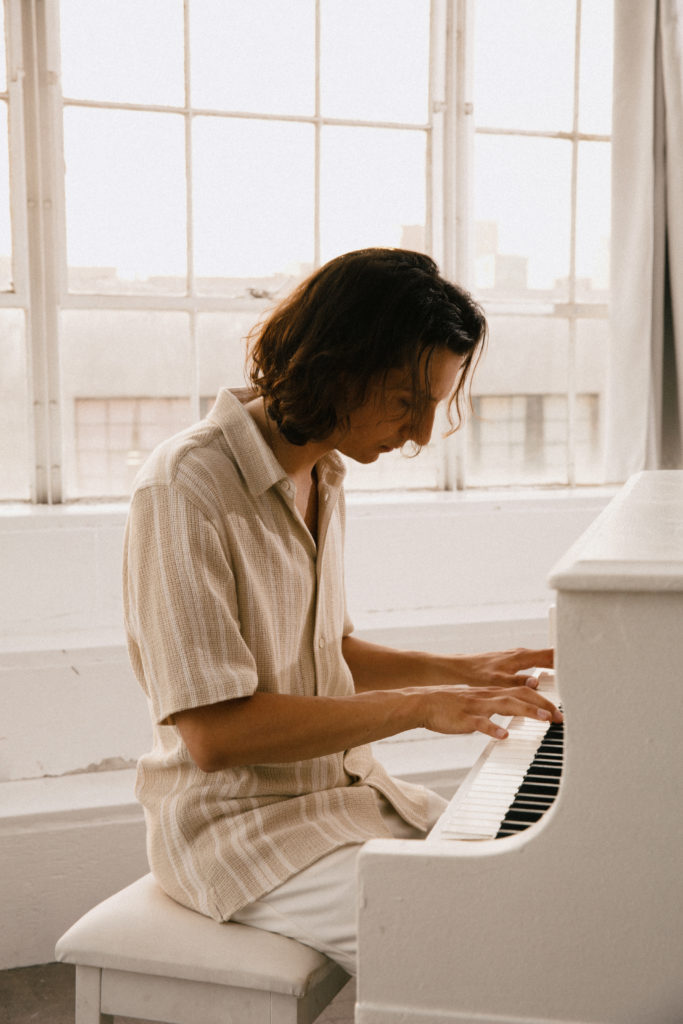 Anson Seabra, a white man with shoulder-length brown hair and a beige shirt, playing a white piano.