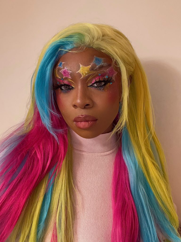 Kryscy Pride Looks - Kryscy is a medium dark brown-skinned person. She is wearing a straight wig with pansexual flag colors: yellow, light blue, and pink. They have bright eye makeup in pansexual flag colors, large stars on her forehead, and thick lashes.