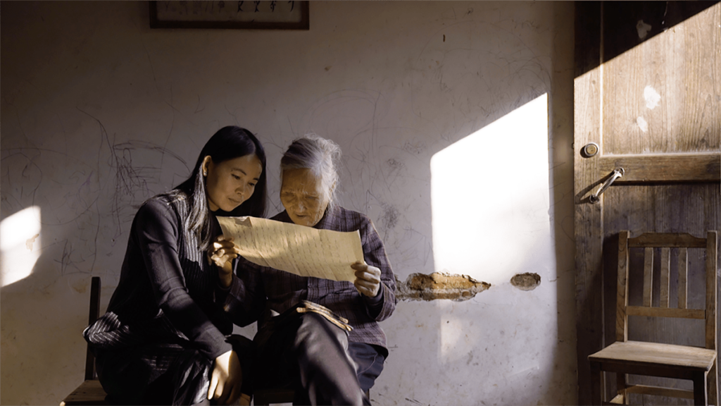 Hu Xin, a young Chinese woman, sits next to He Yan Xin, an older Chinese woman as they both read a paper scroll. Picture is taken from the documentary Hidden Letters.