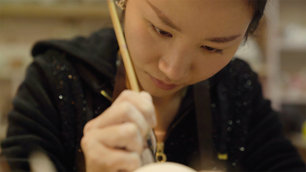 A close up image of Wu Simu, a Chinese woman, writing Nüshu calligraphy on a pot, from the documentary Hidden Letters. She is holding a wooden brush.