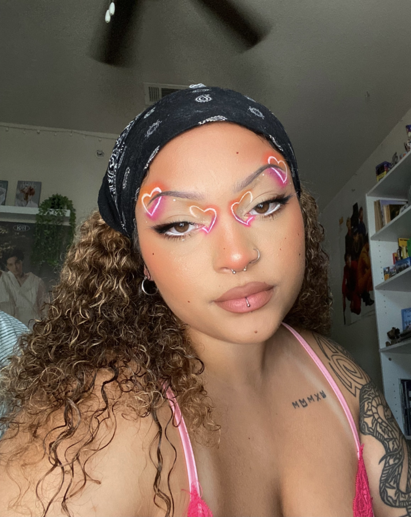 Chanel - EnVi Beauty Team Pride Look. Chanel is a light brown-skinned person with curly light brown hair. She has white hearts, made with eyeliners, with glowing colors of the lesbian flag: orange and pink. They have piercings on their nose and lower lip.