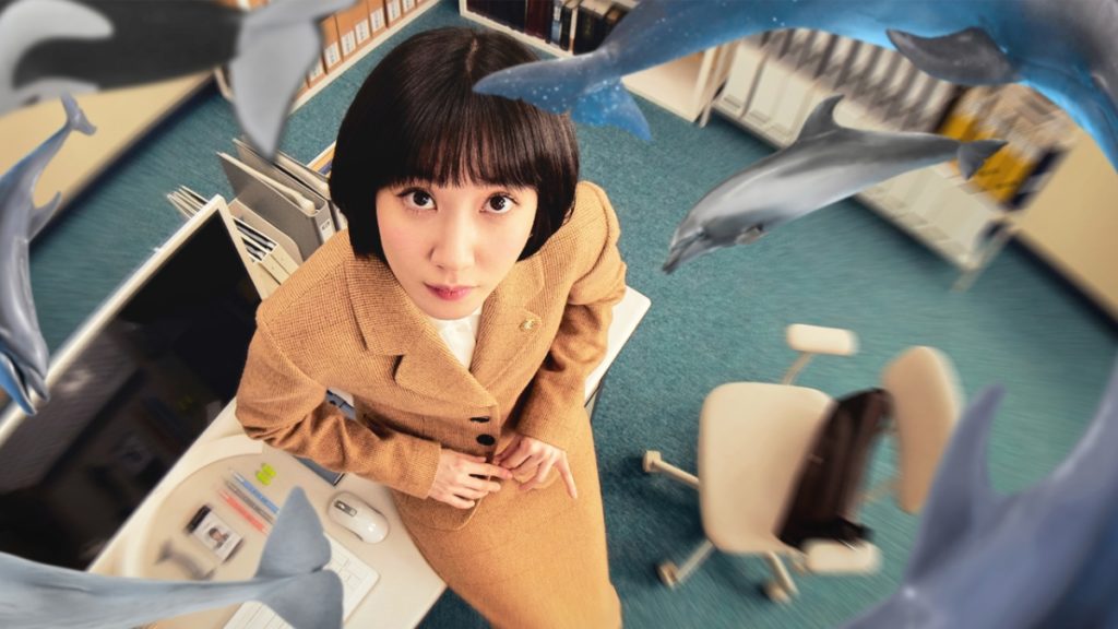 Woo Young-woo, the protagonist of Extraordinary Attorney Woo, looking upwards. She has short black hair with bangs and wears a camel brown suit and skirt. She is surrounded by floating dolphins and whales.