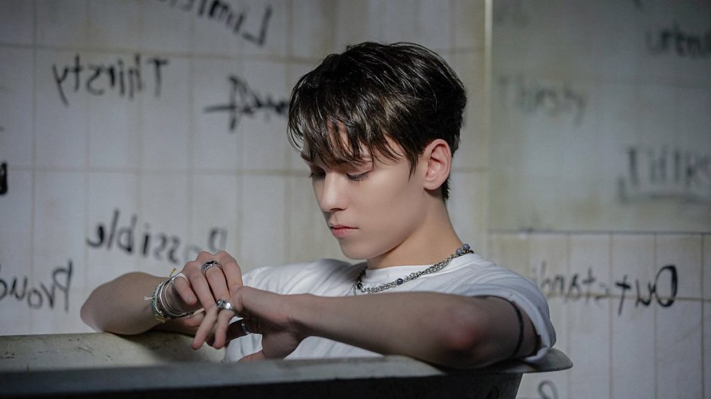 Vernon of SEVENTEEN sitting in a bathtub, surrounded by walls filled with graffiti here and there. His look is emo.