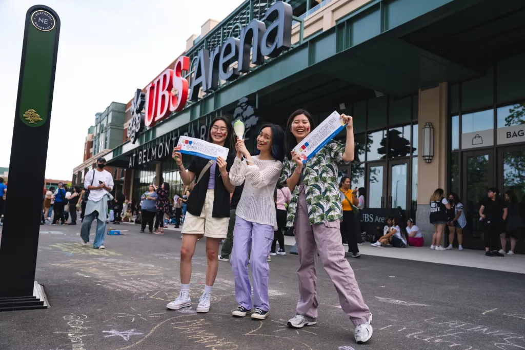 Three MAMAMOO fans posing in front of the UBS Arena, showing their banners and group light stick.