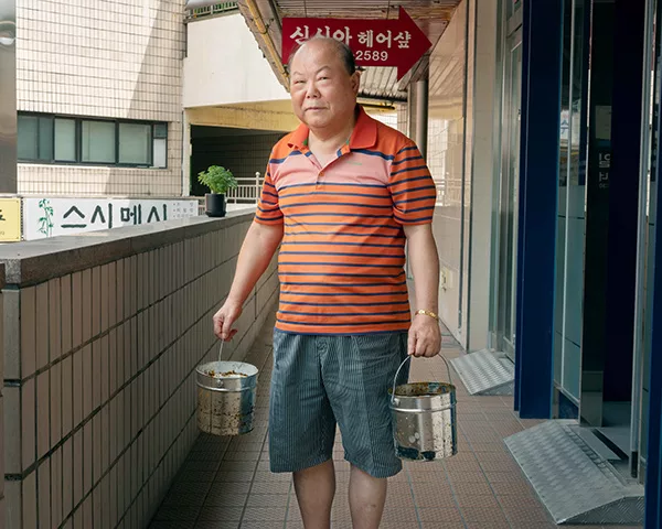 An elderly Korean man holding two metallic buckets.