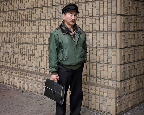 An elderly man standing while holding a leather briefcase. He has a black newsboy cap on.