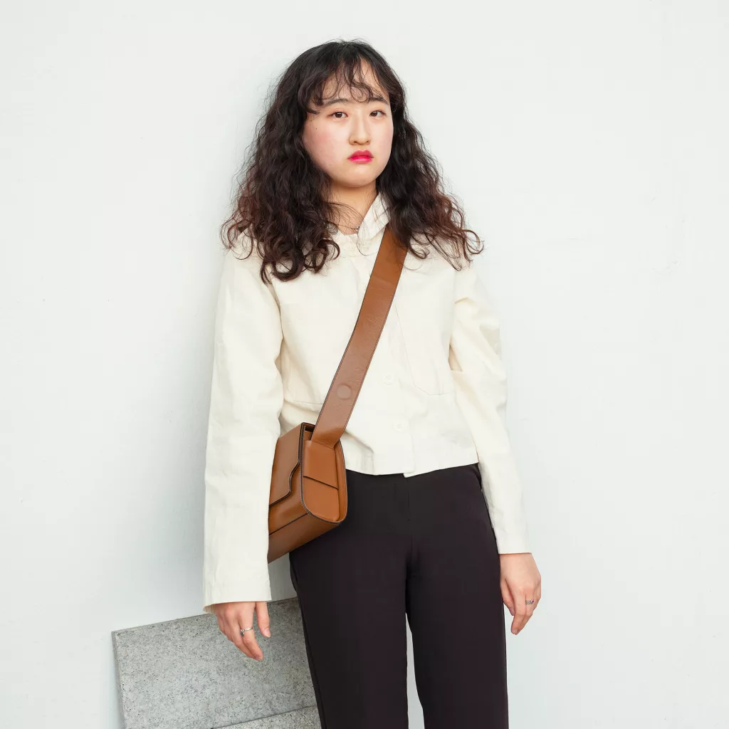 Haneul Lee - A woman with wavy hair, long-sleeved white blouse, and black pants. She has a brown leather sling bag across her body.