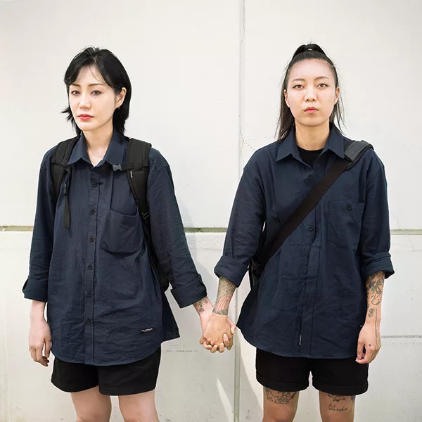Two women holding hands. They are both wearing loose dark gray collared shirts and dark shorts.