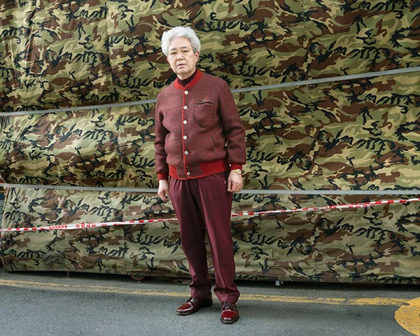 An elderly Korean man standing. He has a mostly burgundy tweed outerwear on and matching trousers.