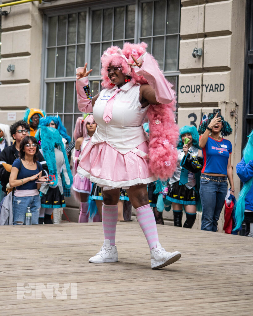 A competitor wears an Alternative version of Hatsune Miku, dressed in pink with a furry afro fixed into two pigtails. This competitor holds hands in a peace sign.