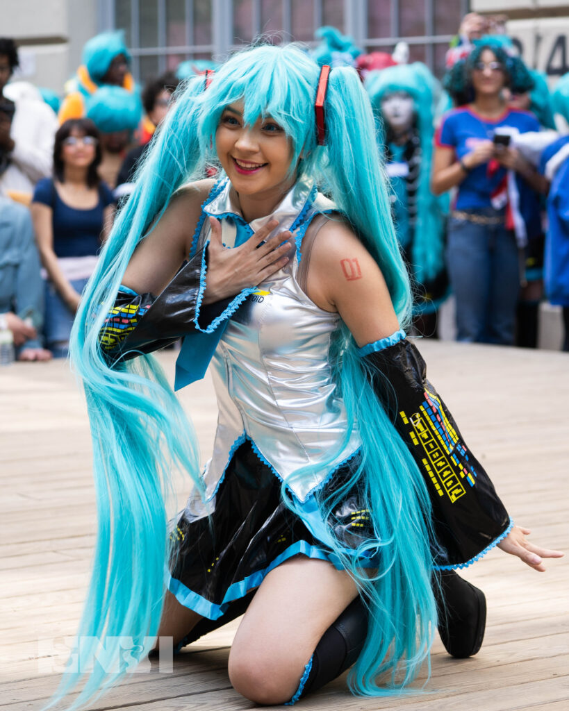Pearlhime is performing to a packed crowd of fans at Japan Village. Wearing a silver sleeveless top with a black skirt, and boots, she smiles while kneeling.