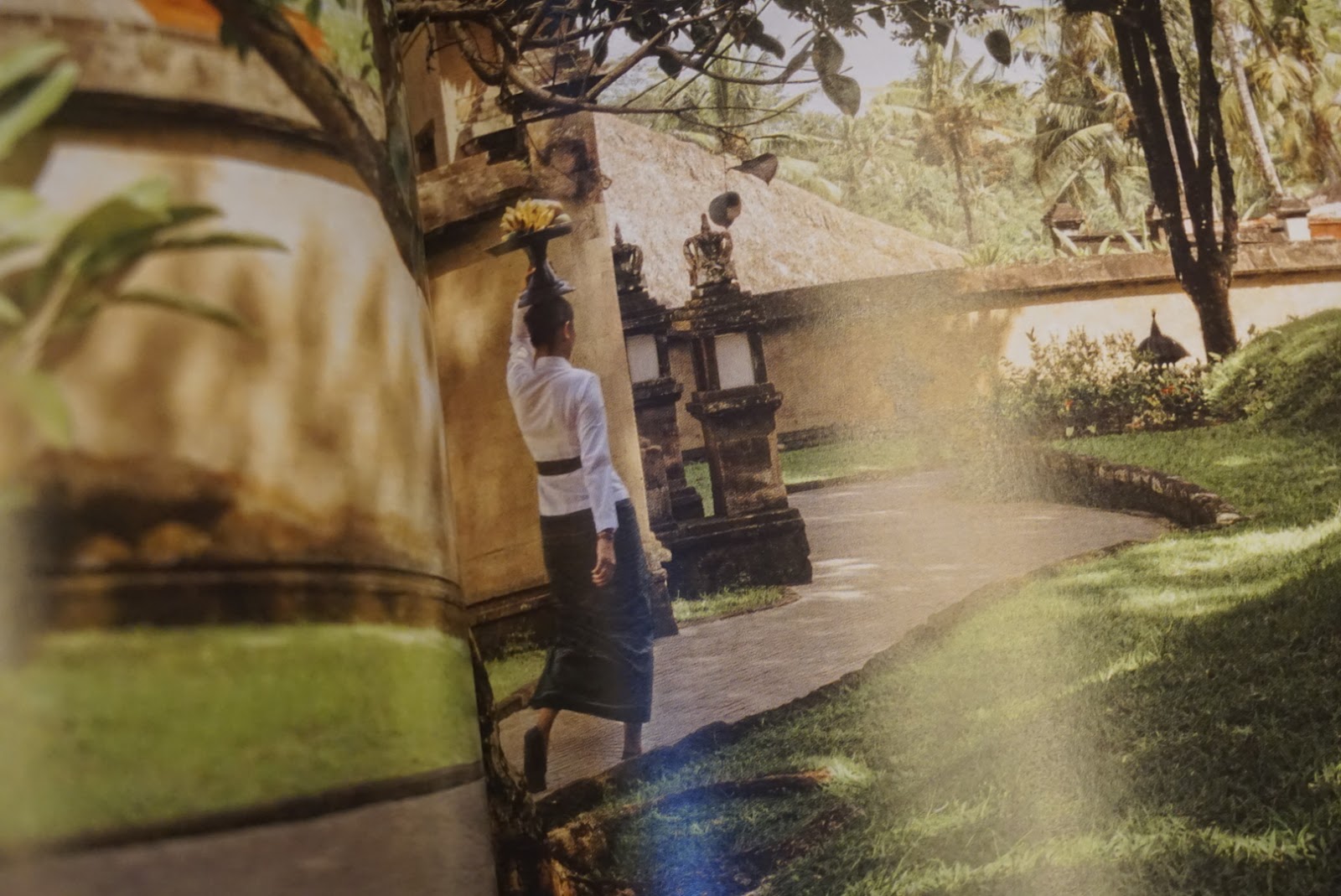 Image from Paras, showcasing a woman dressed in a uniform inspired by traditional Balinese attire holding a tray of fruit over her head walking in a trail of a Balinese hotel